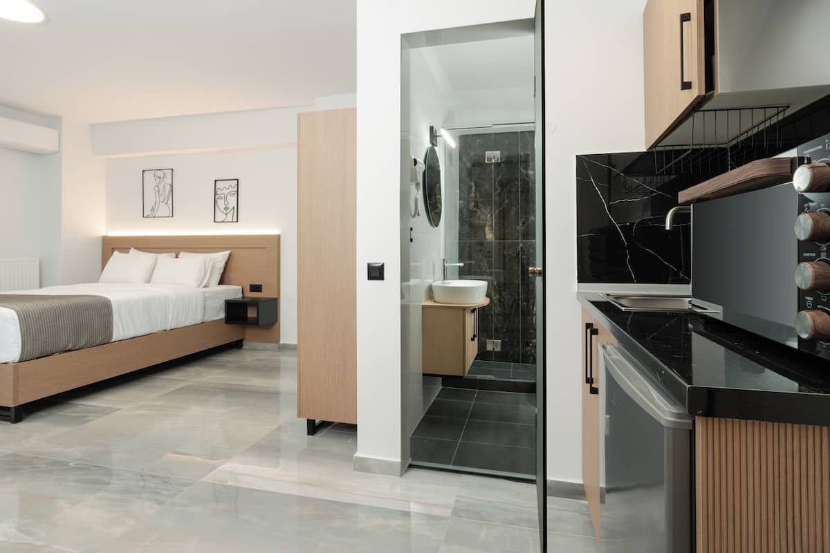 A modern hotel room with a bed, a glass-enclosed shower, a kitchen area, and a bathroom.