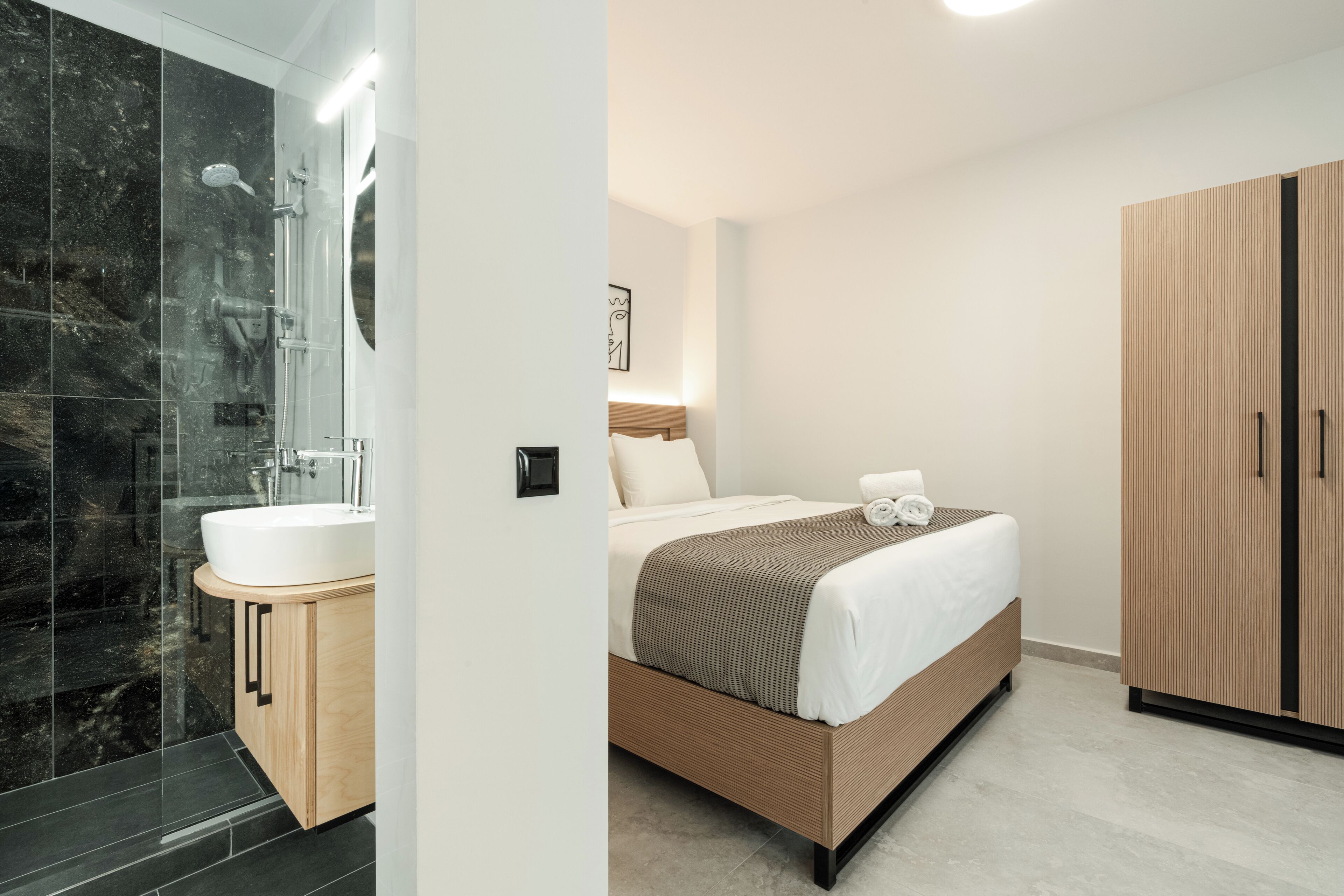 A modern hotel room with a large bed, a wooden wardrobe, a bathroom with a glass shower enclosure, and a white sink.