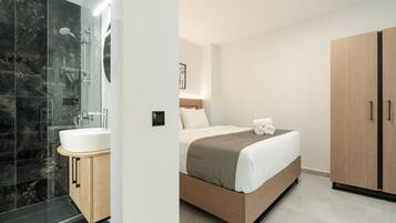 A modern hotel room with a large bed, a wooden wardrobe, a bathroom with a glass shower enclosure, and a white sink.