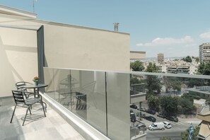 Un balcón con mesa y sillas, una planta en maceta y vistas de un paisaje urbano con edificios y coches estacionados.