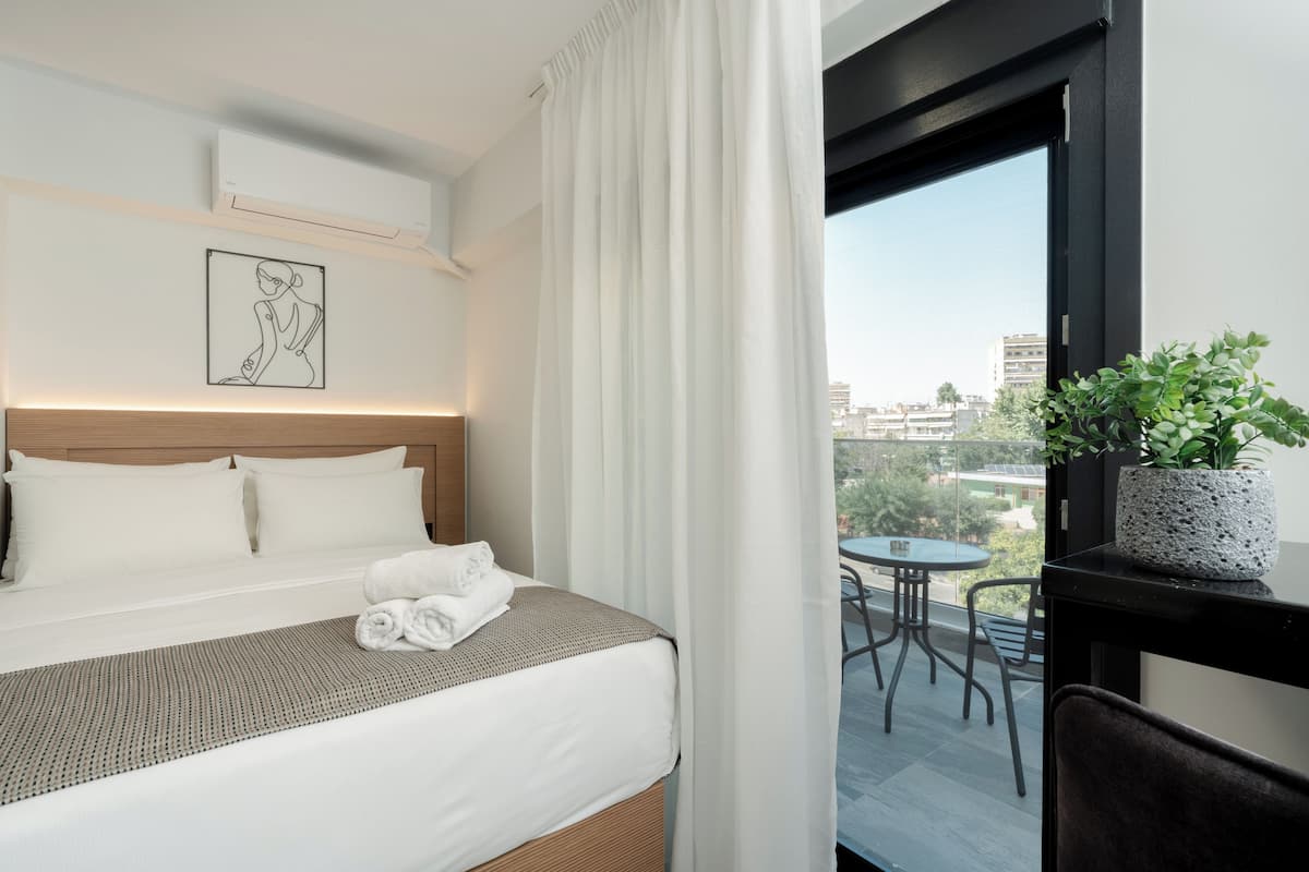 A hotel room with a bed, a framed artwork, a potted plant, and a balcony with a table and chairs.