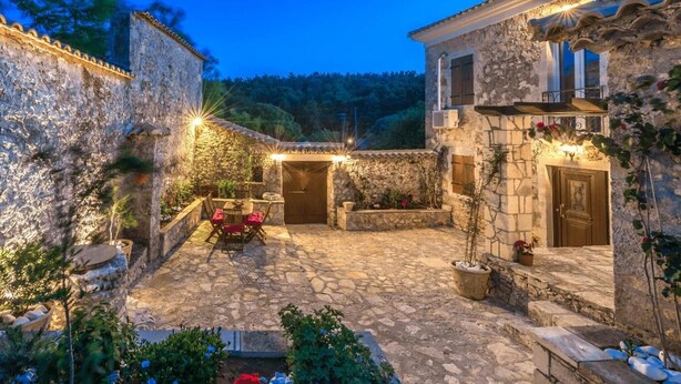 A stone courtyard with a table and chairs, surrounded by buildings with stone walls and wooden doors.