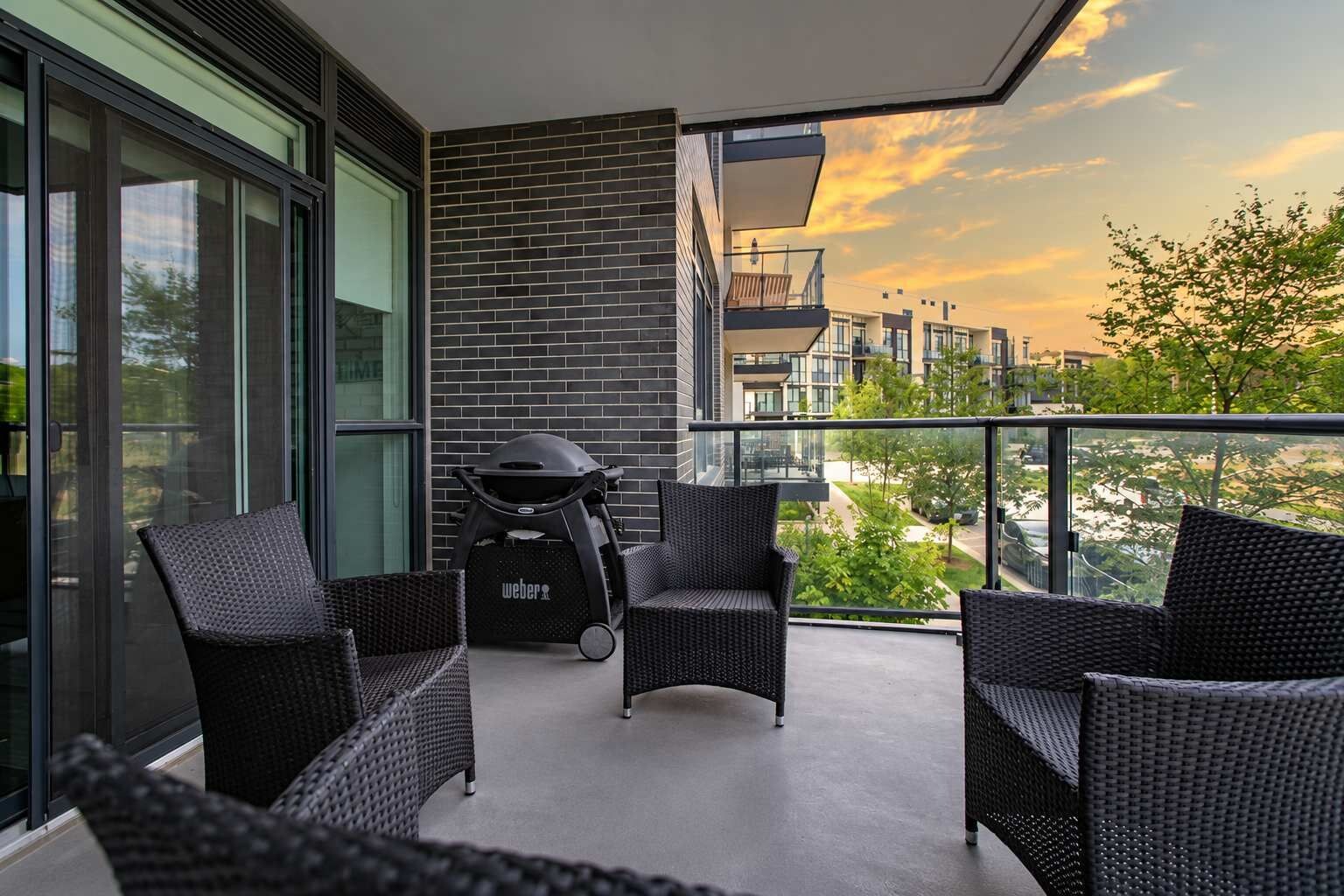 A balcony with wicker furniture, a grill, and a view of buildings and trees.