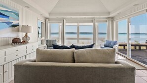 A spacious room with a large window offering a view of the sea, a beige sofa with blue cushions, and a white ceiling with wooden paneling.