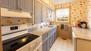 A kitchen with grey cabinets, a white stove, and a microwave. There is a window with a view of trees and a white ceiling light. - Historic PEI Cottage with Modern Comforts, Riverside Views & Family-Friendly Fun (Prince Edward Island)