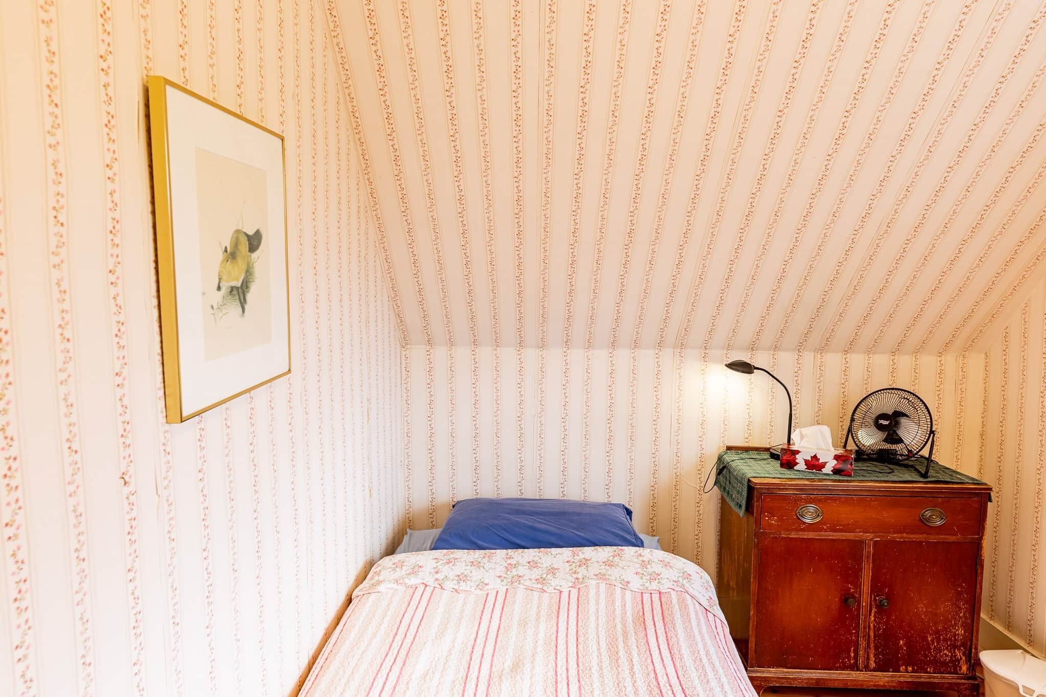 Un dormitorio con papel tapiz a rayas, una cama con colcha estampada, un aparador de madera y un cuadro enmarcado en la pared.