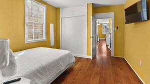 A bedroom with a bed, a TV mounted on the wall, a wardrobe, and a hallway leading to a bathroom. - Beautiful Bywater Retreat with Private Patio (New Orleans)