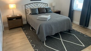 Ein ordentlich eingerichtetes Schlafzimmer mit einem Bett, zwei Nachttischen, einer Lampe, einem gerahmten Bild und einem Fenster mit Vorhängen.