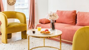 A room with a yellow chair, a pink sofa, a small round table with breakfast items, and a vase with flowers. - Family Suite - Finlay (Paris)