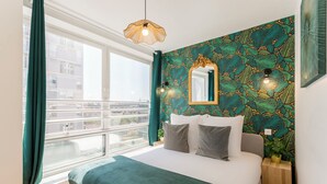 A bedroom with a large bed, a window with curtains, a wall-mounted mirror, and a small bedside table with a plant. - Family Suite - Finlay (Paris)