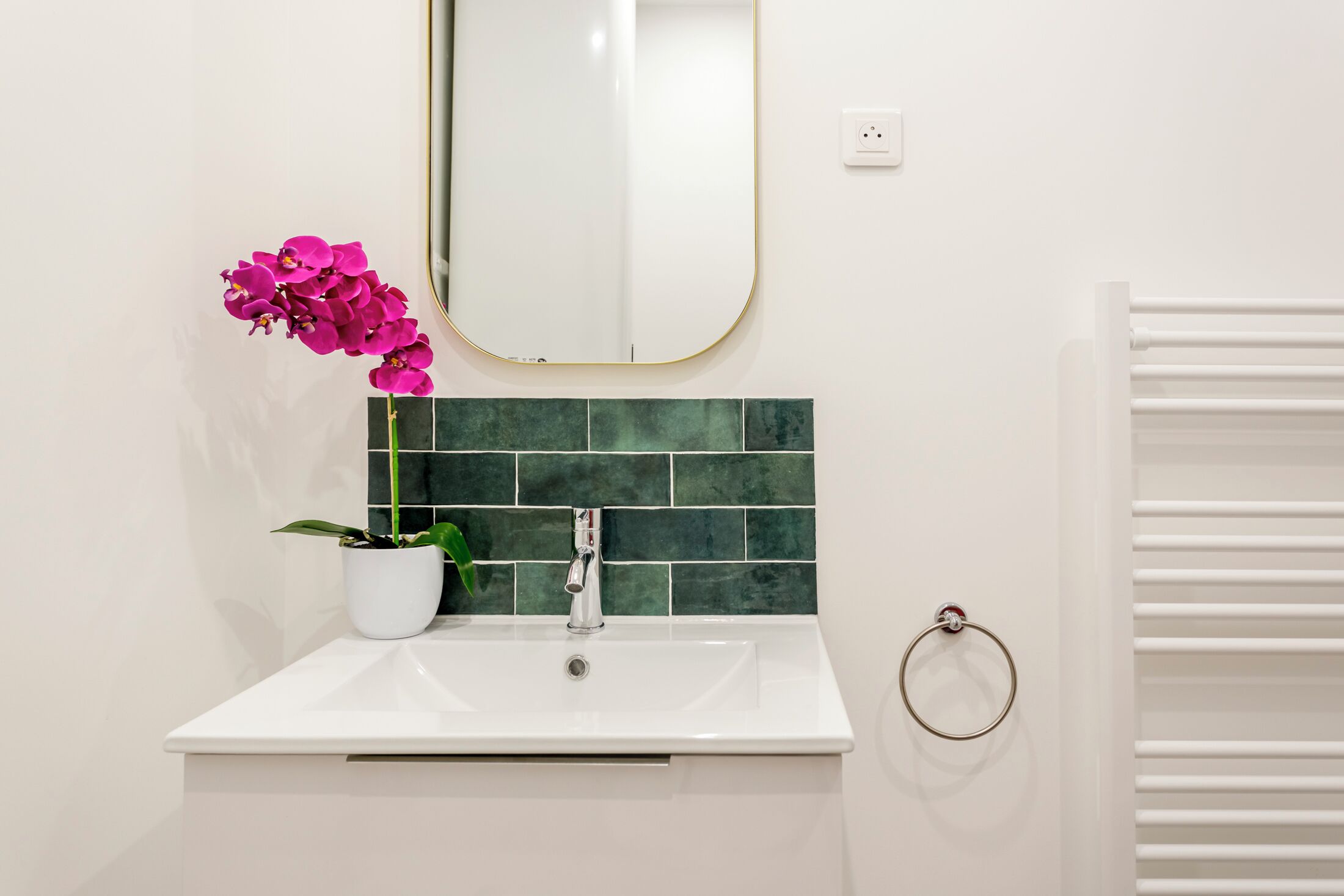 Une salle de bain avec un lavabo blanc, un mur recouvert de carreaux verts, une orchidée en pot et un porte-serviette.