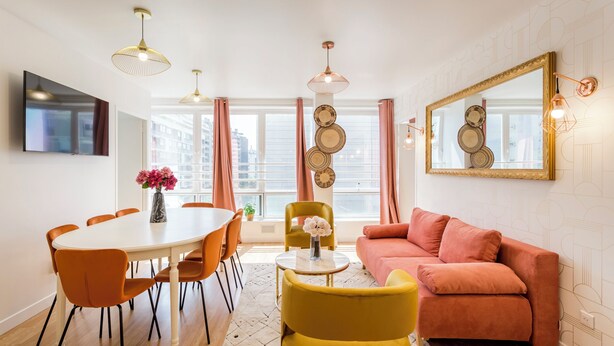 A modern living room with a white dining table, orange chairs, a yellow armchair, a pink sofa, and a large mirror on the wall. - Family Suite - Finlay (Paris)