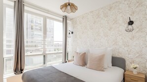A modern bedroom with a large bed, a nightstand, a wall-mounted light, and a window with curtains. - Family Suite - Finlay (Paris)