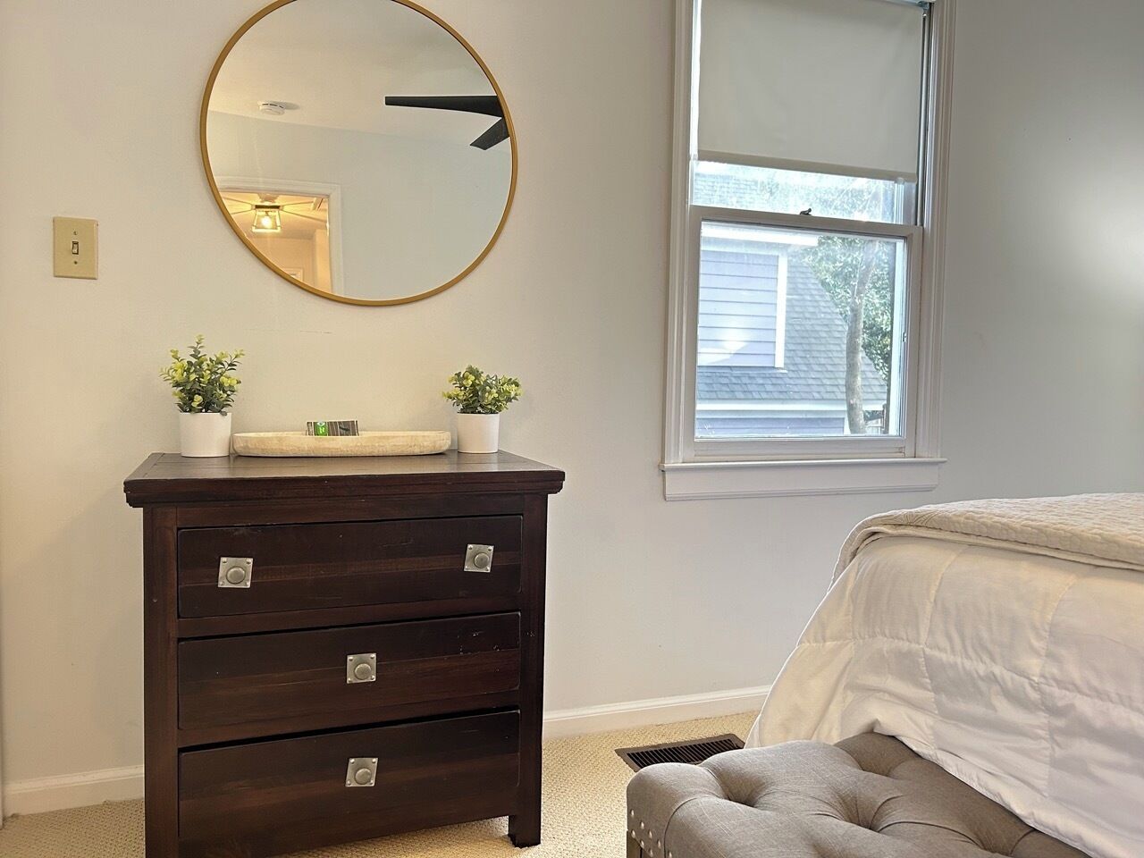 Une chambre avec une commode en bois foncé, un miroir rond, deux plantes en pot, une fenêtre avec des stores et un pouf capitonné.