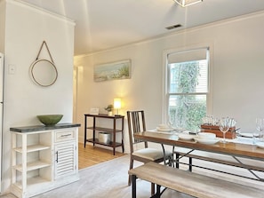 Un coin repas avec une table en bois dressée pour quatre personnes, un banc, une petite armoire blanche et un tableau accroché au mur.