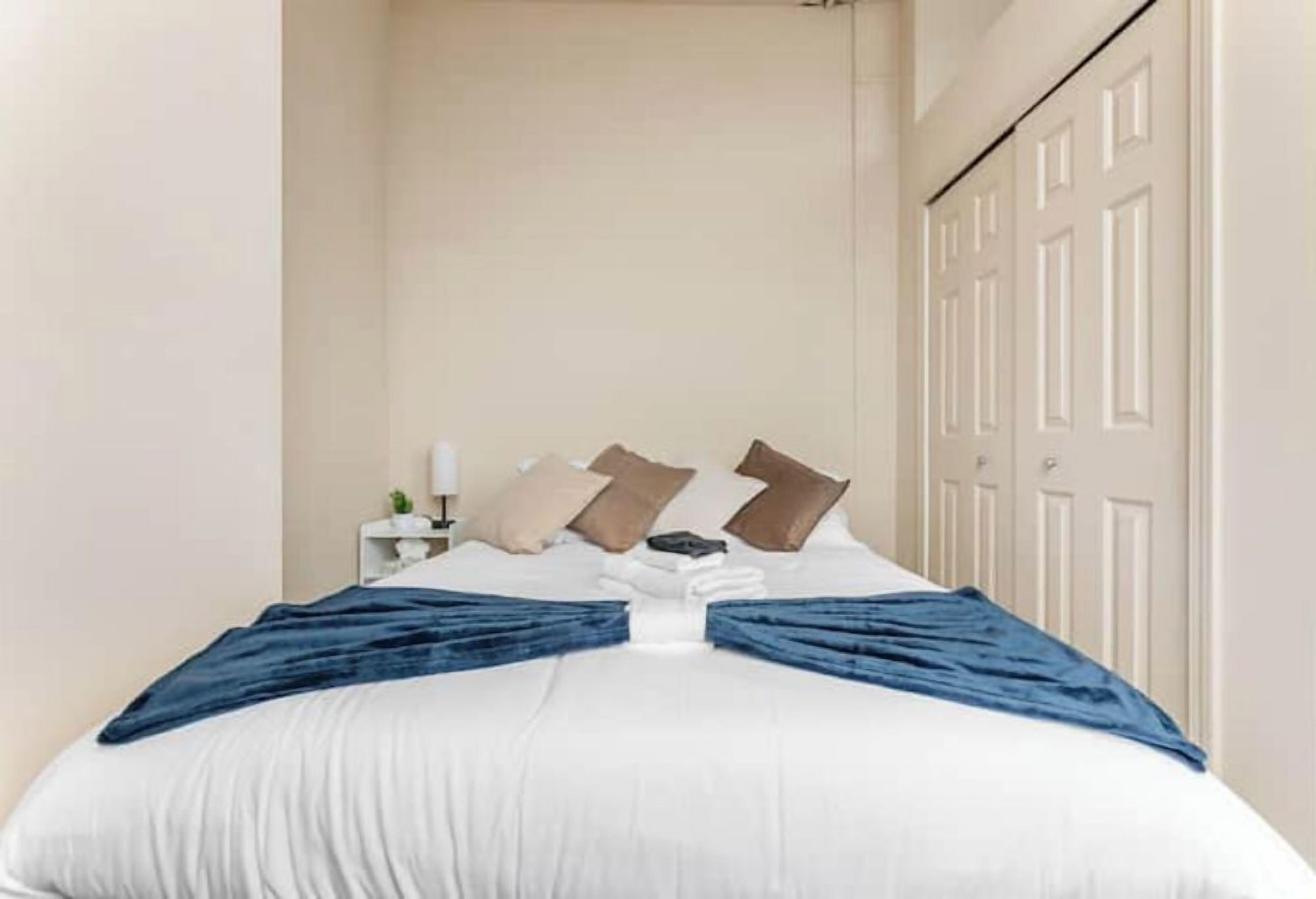 Una cama bien hecha con una colcha blanca y azul, una mesita de noche con una lámpara, y una puerta al fondo.