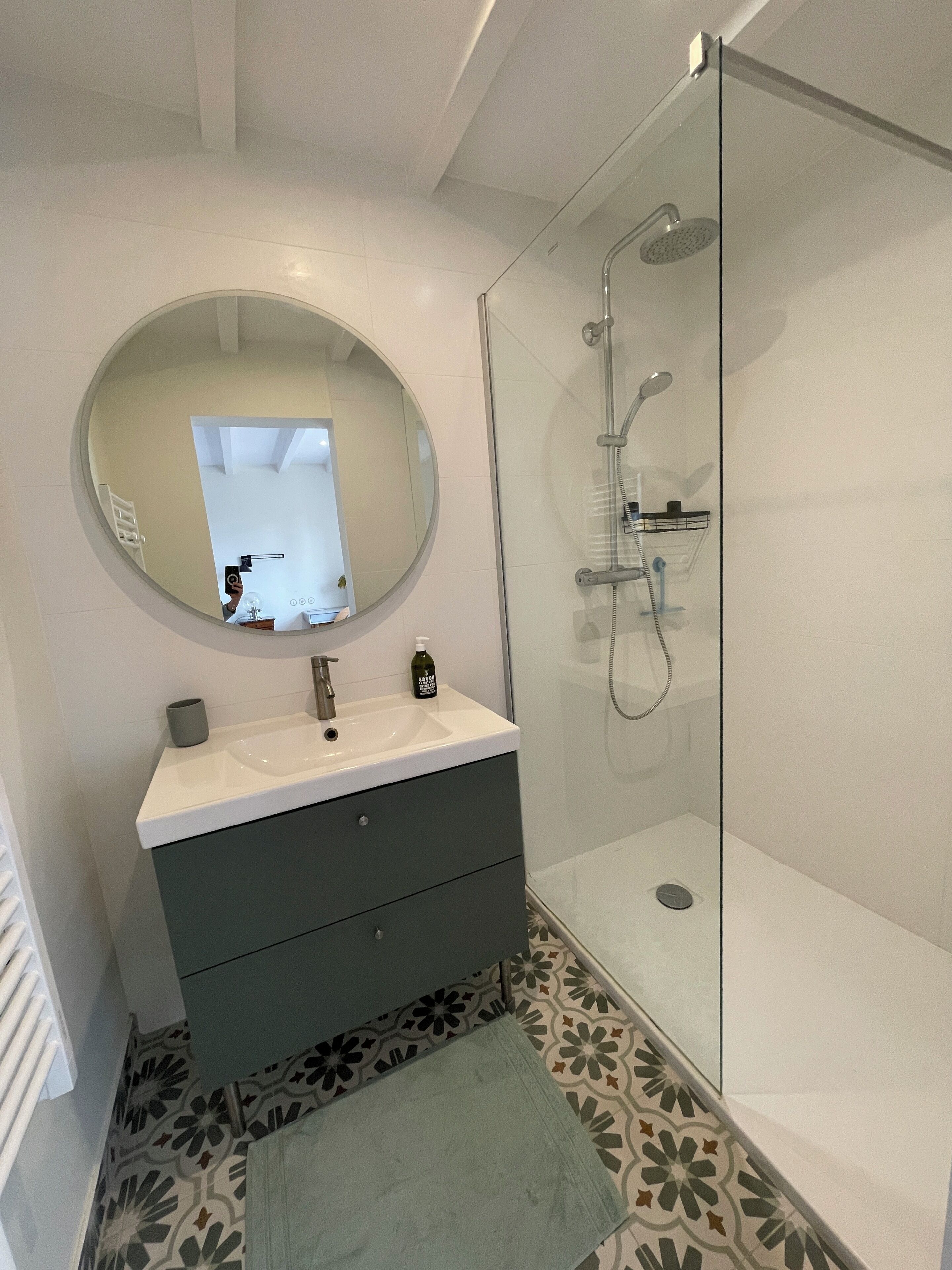 A modern bathroom with a round mirror, a sink, a shower, and patterned floor tiles.
