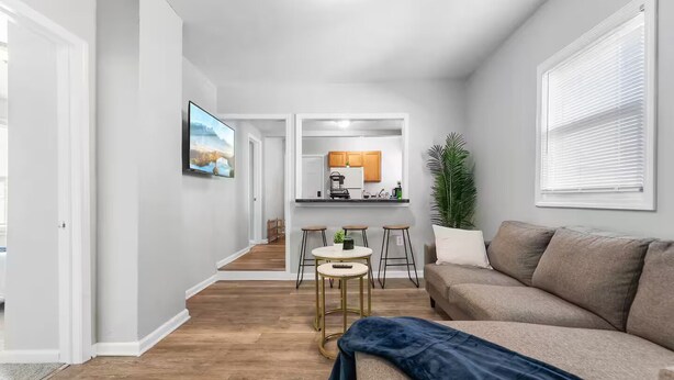 A modern living room with a sofa, a coffee table, and a TV. There is a kitchen area with a countertop and cabinets.
