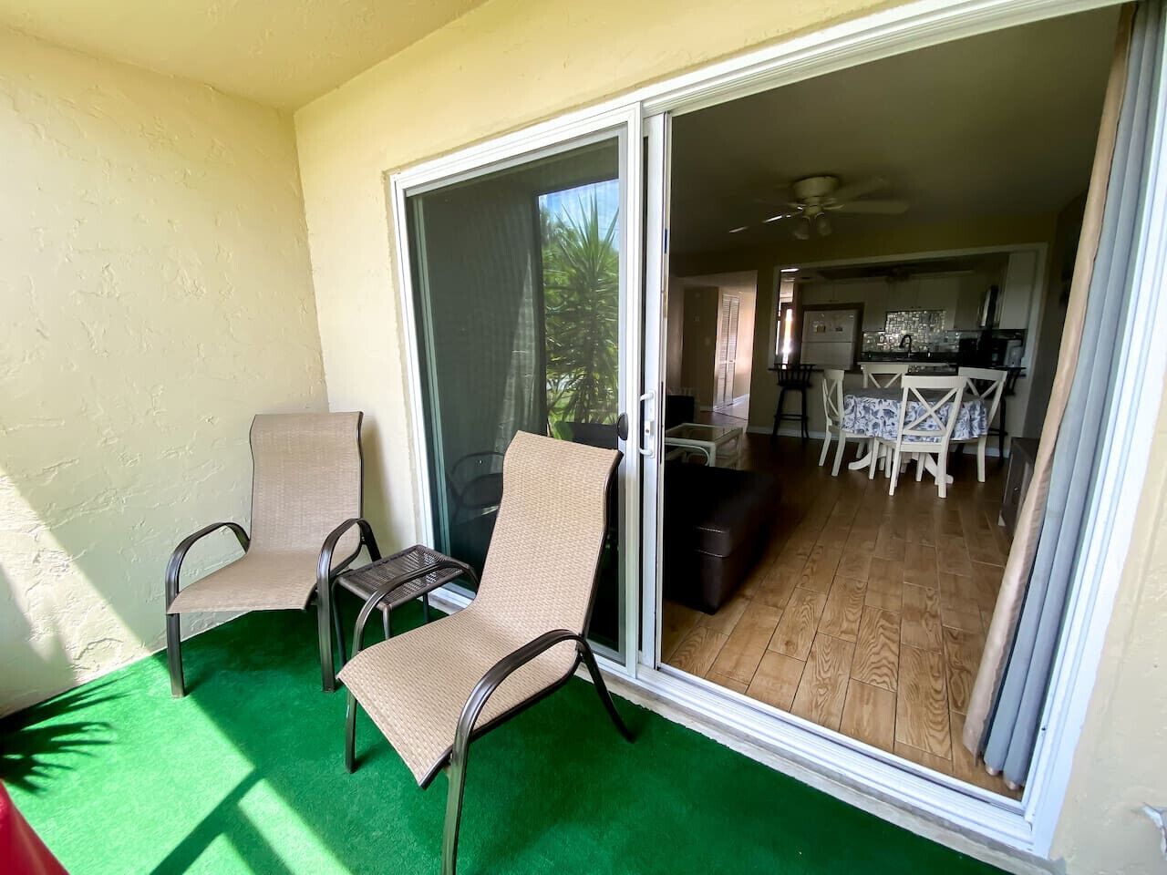 Un balcon avec deux chaises et une table, une porte-fenêtre coulissante donnant sur un coin repas équipé d’une table et de chaises, et un ventilateur de plafond.