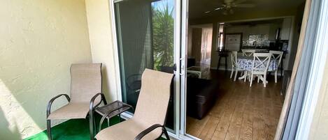 Un balcon avec deux chaises et une table, une porte-fenêtre coulissante donnant sur un coin repas équipé d’une table et de chaises, et un ventilateur de plafond.
