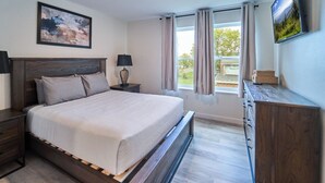 Une chambre à coucher avec un grand lit, une tête de lit en bois, deux tables de chevet avec des lampes, une commode, un téléviseur fixé au mur et une fenêtre avec des rideaux.