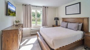 Quarto com cama grande, mesa de cabeceira de madeira, mesa lateral, TV fixada na parede e janela com cortinas.