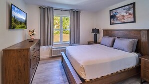 Un dormitorio con una cama de madera, una mesita de noche, un aparador, un televisor en la pared y una ventana con cortinas.