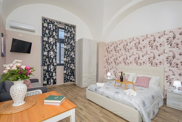 Une chambre à coucher avec un lit, des tables de chevet, une télévision, une fenêtre avec des rideaux et un vase rempli de fleurs.