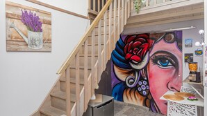 A staircase with a wooden handrail, a painting of a watering can with flowers, and a mural of a woman's face.