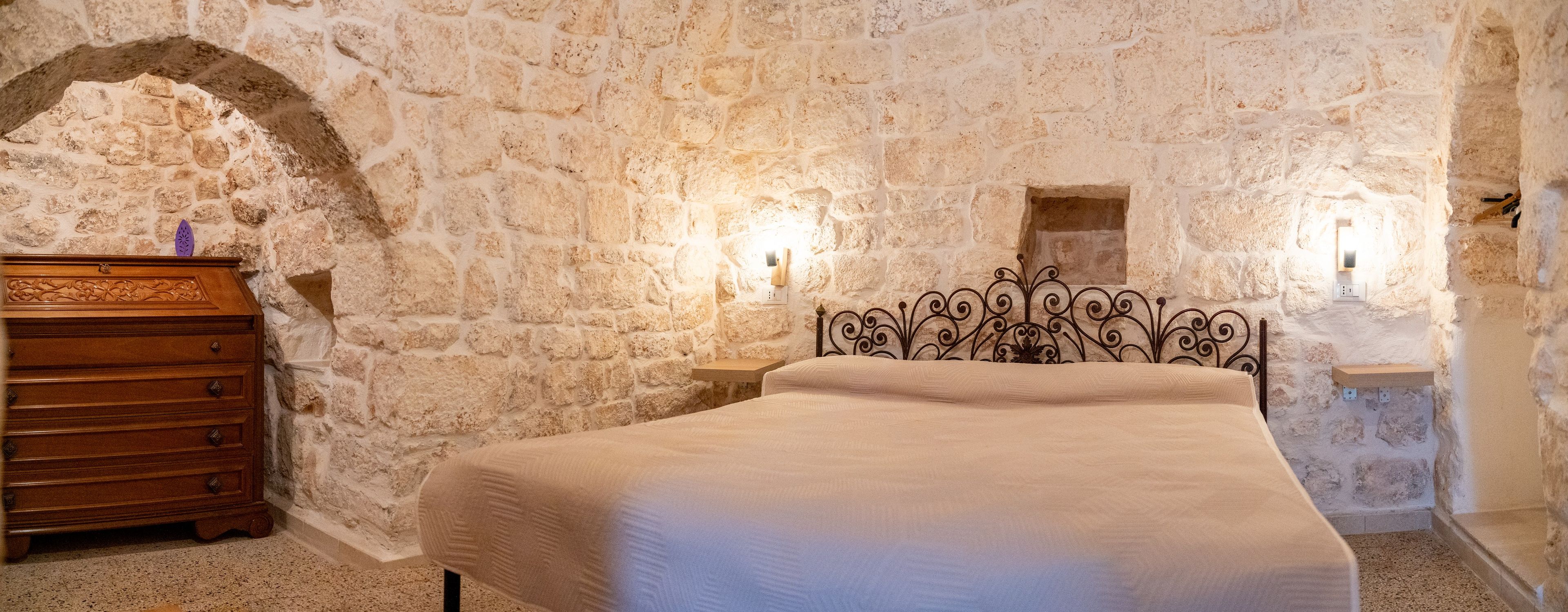 A bedroom with a stone wall, a wooden dresser, a metal bed frame, and two wall-mounted lights.