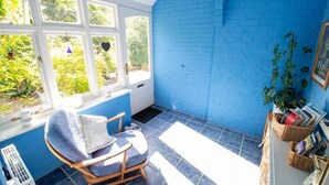 Une pièce avec un mur en briques bleues, une fenêtre donnant sur un paysage verdoyant, un fauteuil avec un coussin et une bibliothèque remplie de livres et agrémentée d’une plante.