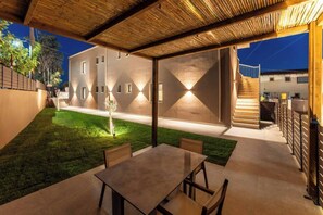 Un espace extérieur aménagé en terrasse, avec une table et des chaises, une pelouse verdoyante et un bâtiment au design moderne, doté d’un éclairage chaleureux.