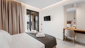 A modern hotel room with a large bed, a desk with a chair, a flat-screen TV, and a sliding glass door leading to a balcony.