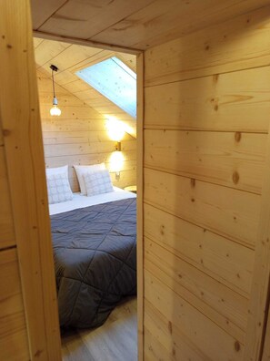 Um quarto de madeira com cama, claraboia e luminária na parede.