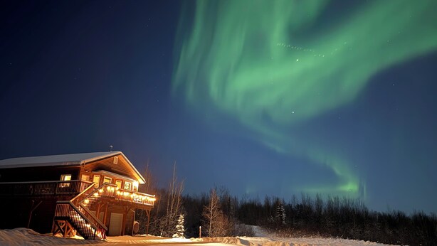 A lodge with a wooden exterior and a balcony, surrounded by snow and trees, under a night sky with the Northern Lights. - Modern 4-bed home, 10 min from town. Deck with Aurora and mountain views, pond, updated kitchen, master suite with jet tub, radiant floors, fast Wi-Fi. Open concept living and dining. Beautifully renovated inside—cozy, private, and peaceful. (Fairbanks)