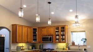 A modern kitchen with wooden cabinets, a central island with bar stools, pendant lighting, and a built-in microwave. - Modern 4-bed home, 10 min from town. Deck with Aurora and mountain views, pond, updated kitchen, master suite with jet tub, radiant floors, fast Wi-Fi. Open concept living and dining. Beautifully renovated inside—cozy, private, and peaceful. (Fairbanks)