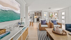 A modern living room with a large flat-screen TV, wicker furniture, and a kitchen in the background.