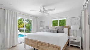 A bedroom with a large bed, a ceiling fan, a nightstand, and a view of a swimming pool.