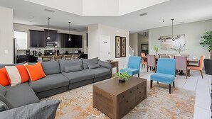 A modern living room with a grey sectional sofa, a coffee table, and a dining area with blue chairs. - 5BR w/ South Pool, Game Room – Storey Lake (Kissimmee)