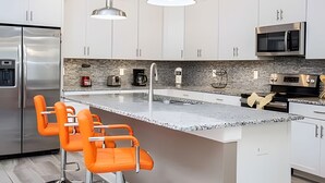 A modern kitchen with white cabinets, a large island with orange bar stools, stainless steel appliances, and pendant lighting. - 6BR Townhome w/ Pool – 10 Min to Disney (Kissimmee)