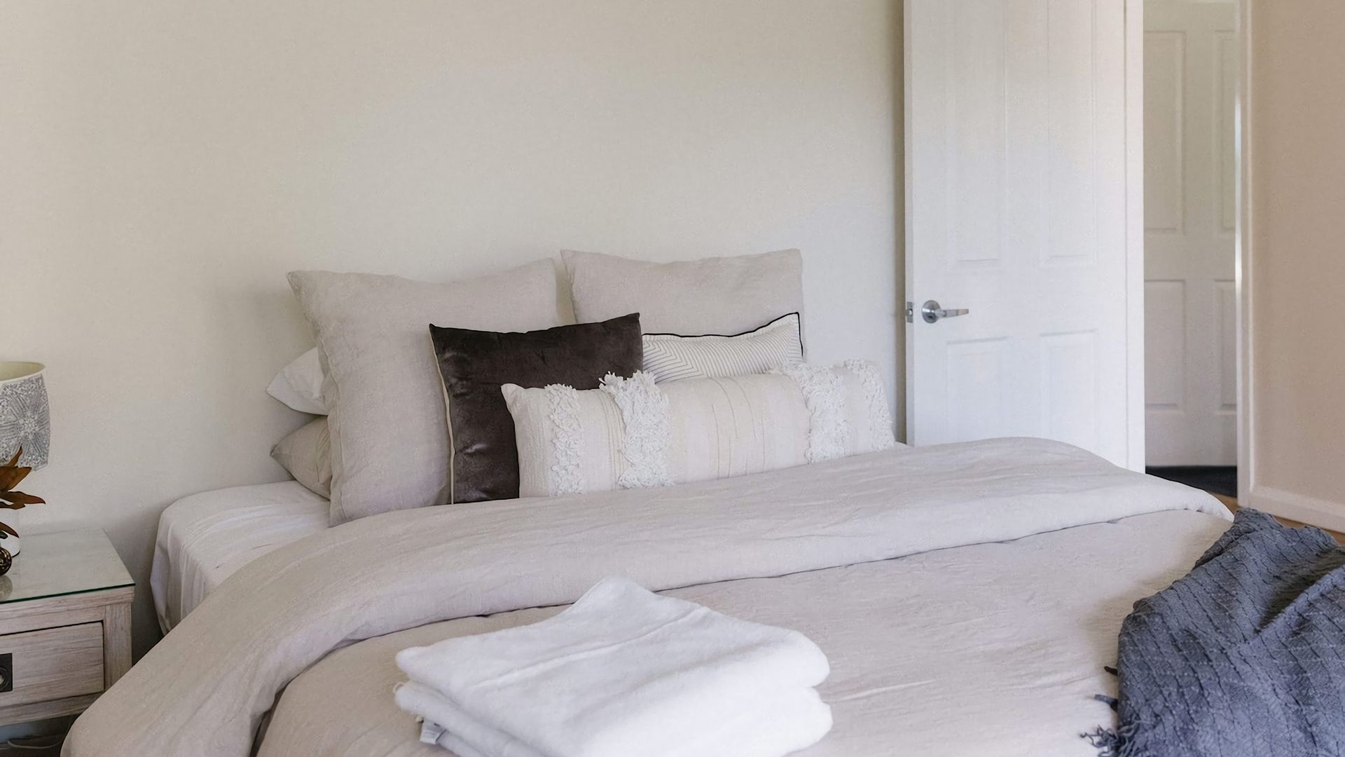 A neatly made bed with a white duvet, several pillows, and a folded towel on a wooden bedside table.