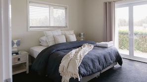 A bedroom with a bed, a nightstand, a window with blinds, and a view of greenery outside.