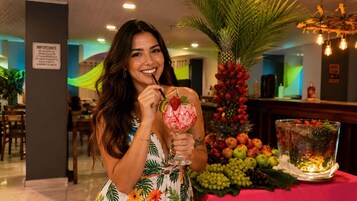 Una persona sostiene una copa de cóctel con una bebida tropical, de pie en una habitación con una mesa llena de frutas y una planta decorativa.