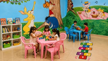 Zona de juegos infantiles con murales de dibujos animados, una estantería con libros para niños y un juego educativo para aprender números.