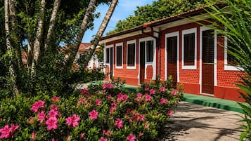 Un edificio de ladrillo rojo con marcos de ventana blancos y techo verde, rodeado de exuberante follaje y flores rosadas.
