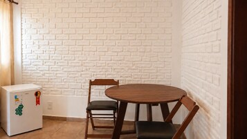 Habitación compacta con pared de ladrillo blanco, un refrigerador blanco pequeño, una mesa de madera con dos sillas y una puerta de madera.