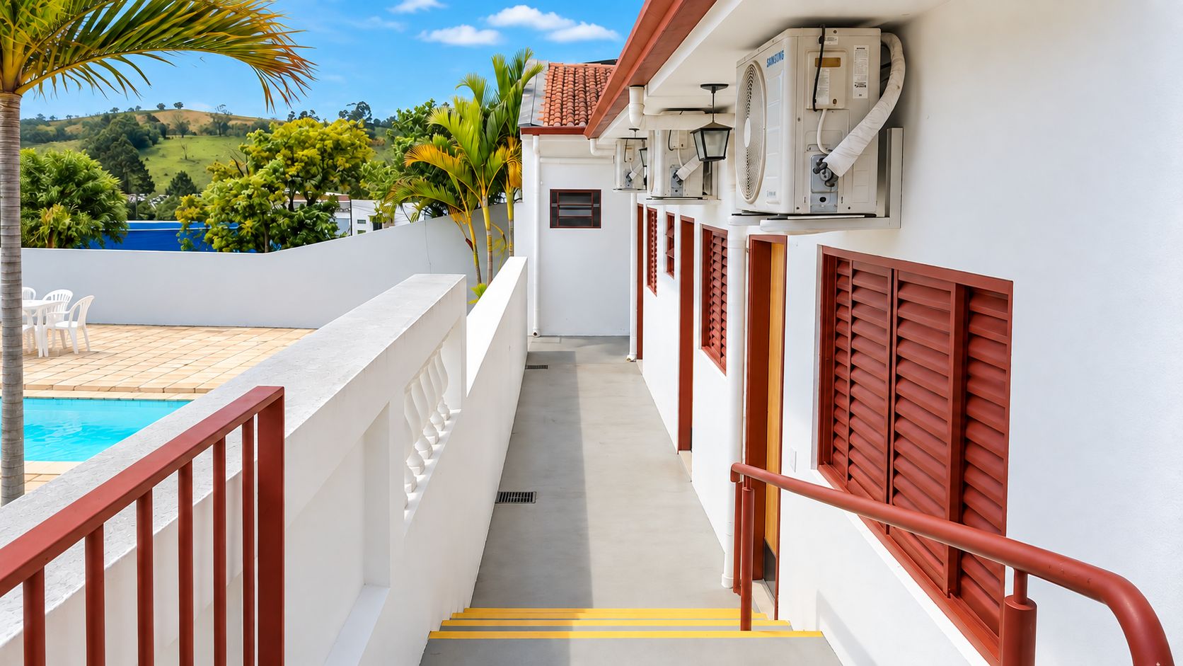 A poolside corridor with a view of a pool and palm trees, leading to a building with air conditioning units and red shutters.