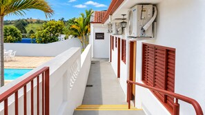 A poolside corridor with a view of a pool and palm trees, leading to a building with air conditioning units and red shutters.