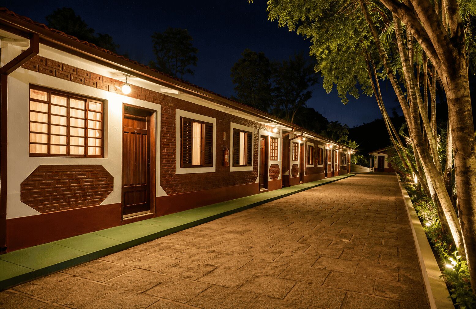 Uma fileira de edifícios de tijolo com várias janelas e portas, um passeio pavimentado e árvores ao fundo.
