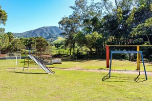 A playground with a slide and swings, surrounded by grass and trees, with a mountain in the background. - Hotel Fazenda Águas de Lindóia (Águas de Lindóia)